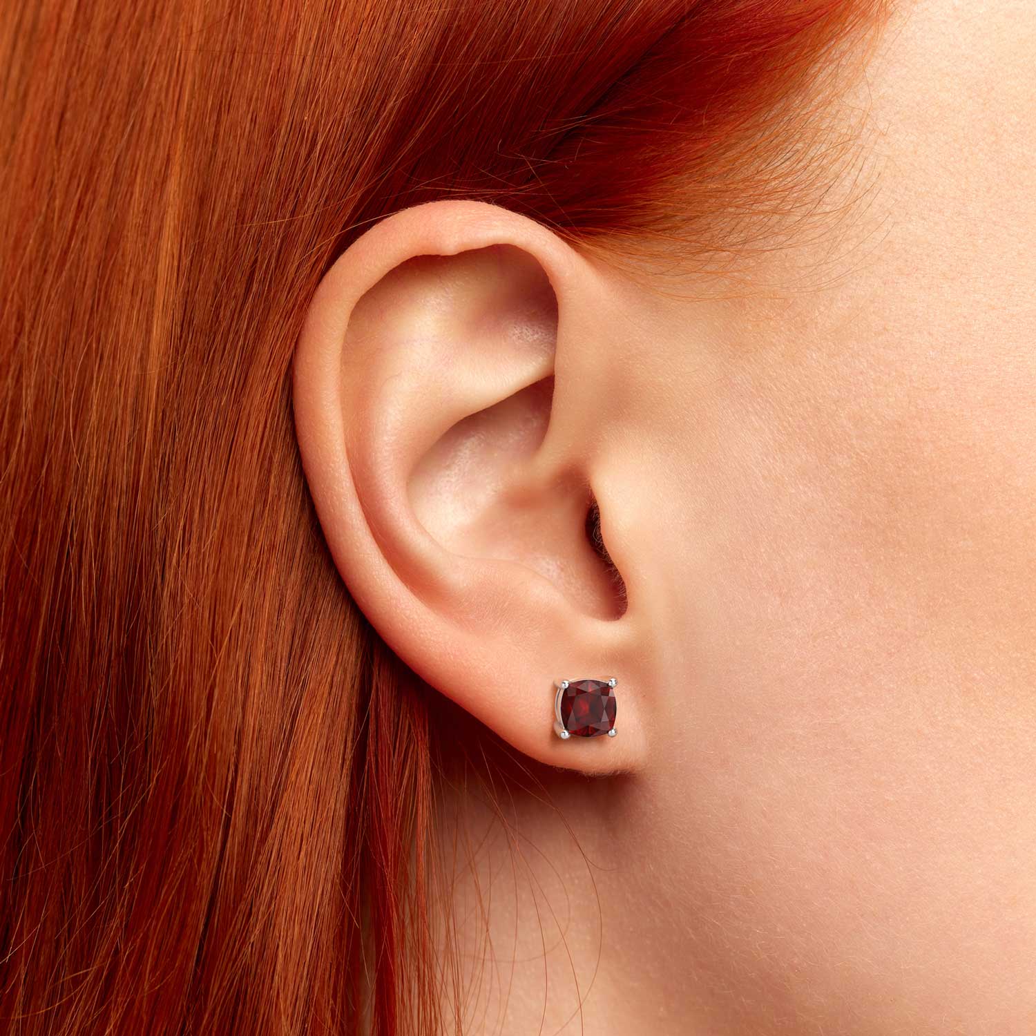 Close-up of a woman’s ear wearing a 6×6 mm cushion-cut garnet stud earring in a 925 sterling silver four-prong setting.