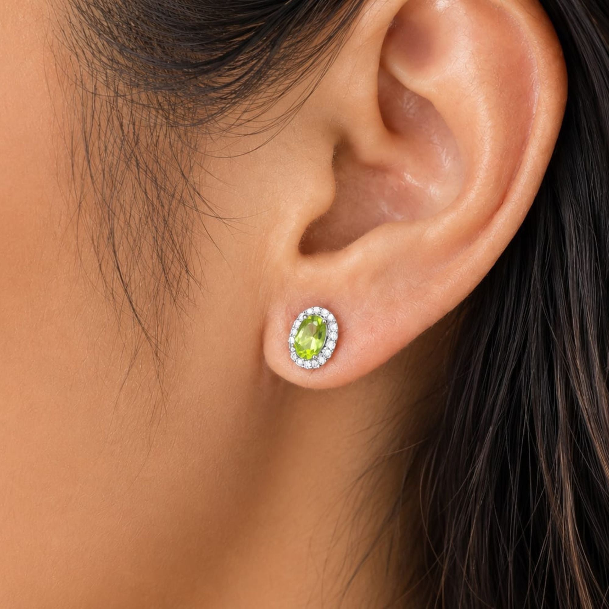 A close-up of a person's ear wearing a small peridot halo oval stud earring with a bright green center stone surrounded by a halo of clear stones.