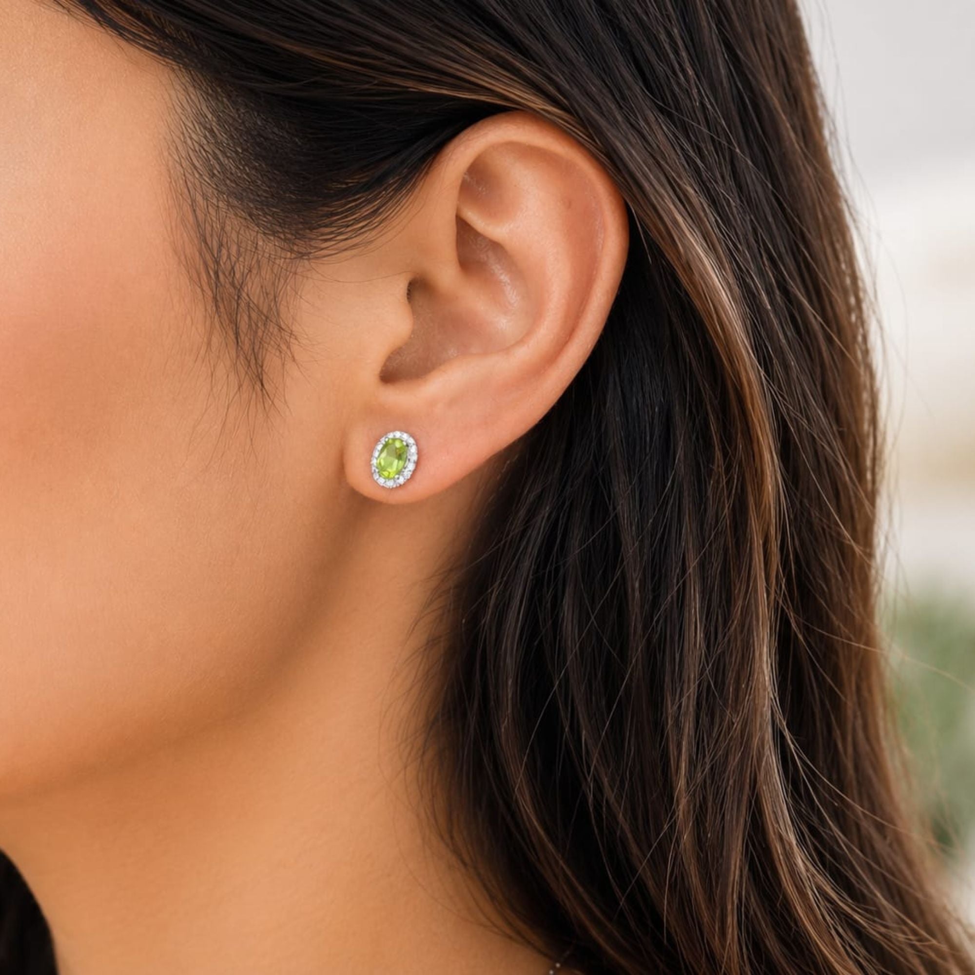 A close-up of a woman's ear wearing a small oval peridot stud earring with a bright green center stone surrounded by a halo of clear stones.
