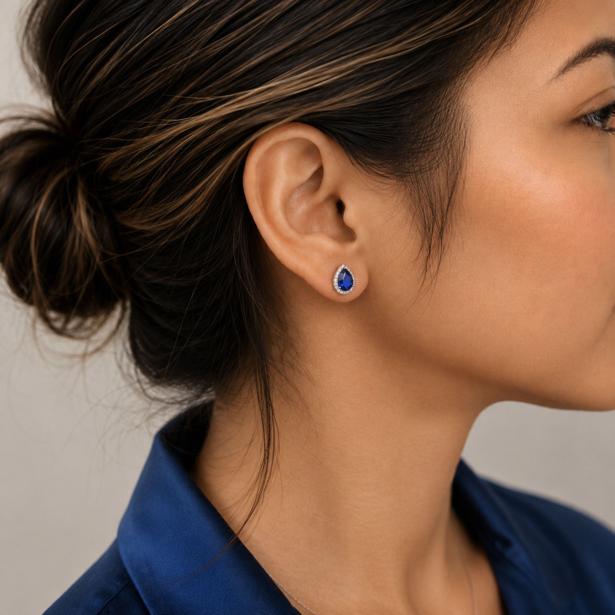 A close-up side profile of a woman wearing a small teardrop-shaped blue sapphire gemstone stud earring with a silver-toned setting.