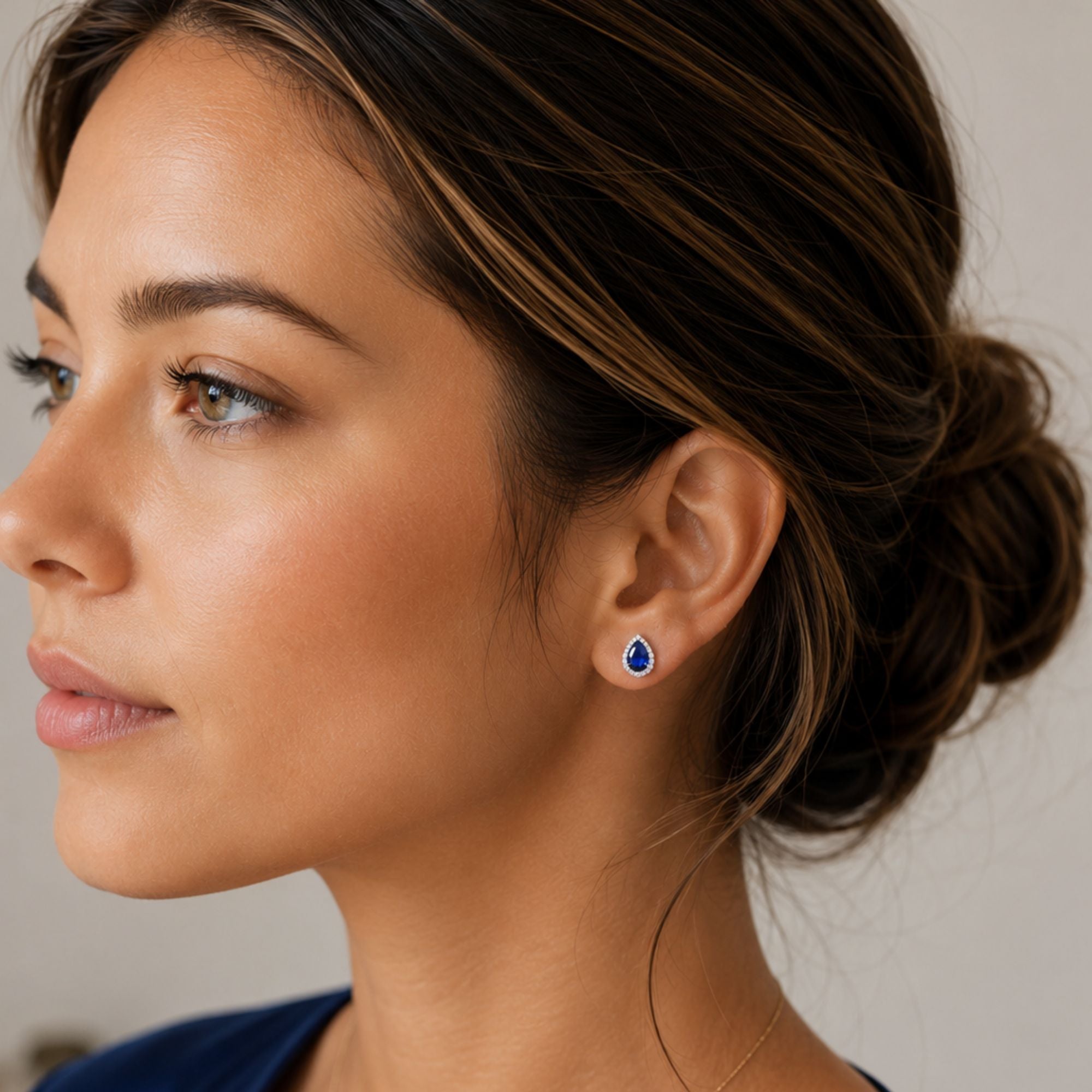 A close-up side profile of a woman with dark hair in a low bun, wearing a small teardrop-shaped blue sapphire gemstone stud earring with a silver setting.