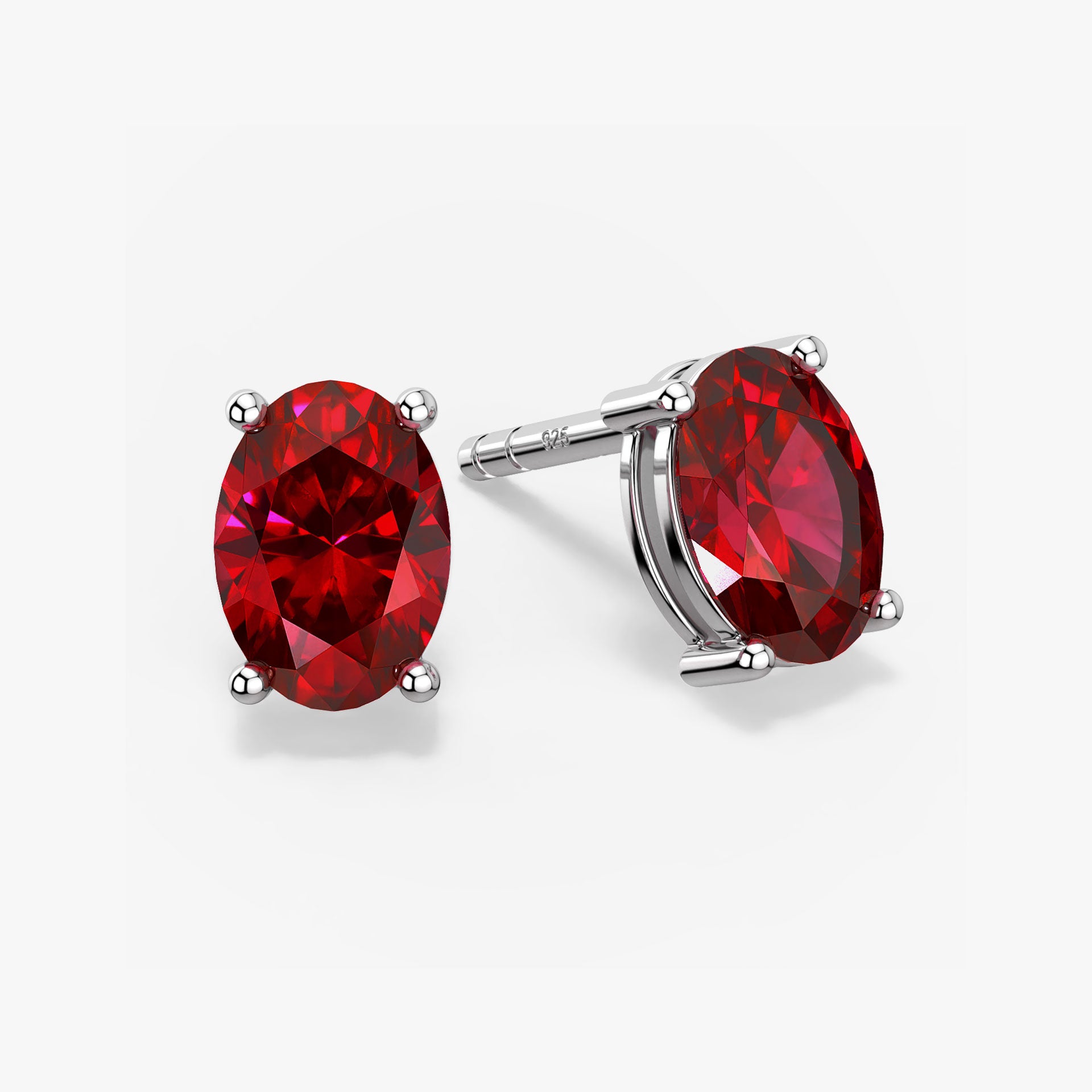 Pair of oval-cut vibrant red ruby stud earrings in a four-prong silver setting with push-back closures, displayed on a white background.