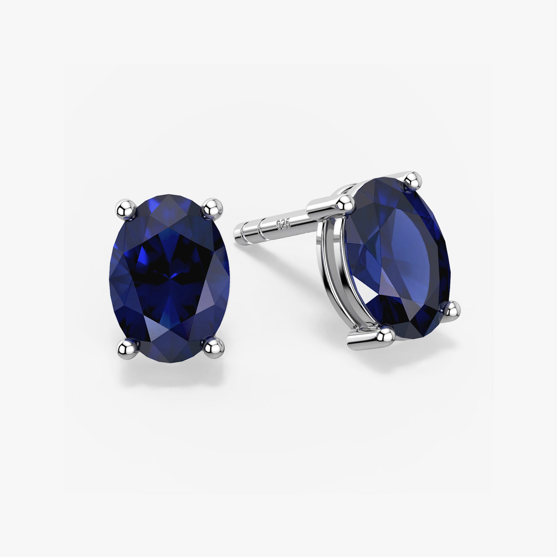 Pair of oval-cut deep blue sapphire stud earrings in a four-prong silver setting with push-back closures, displayed on a white background