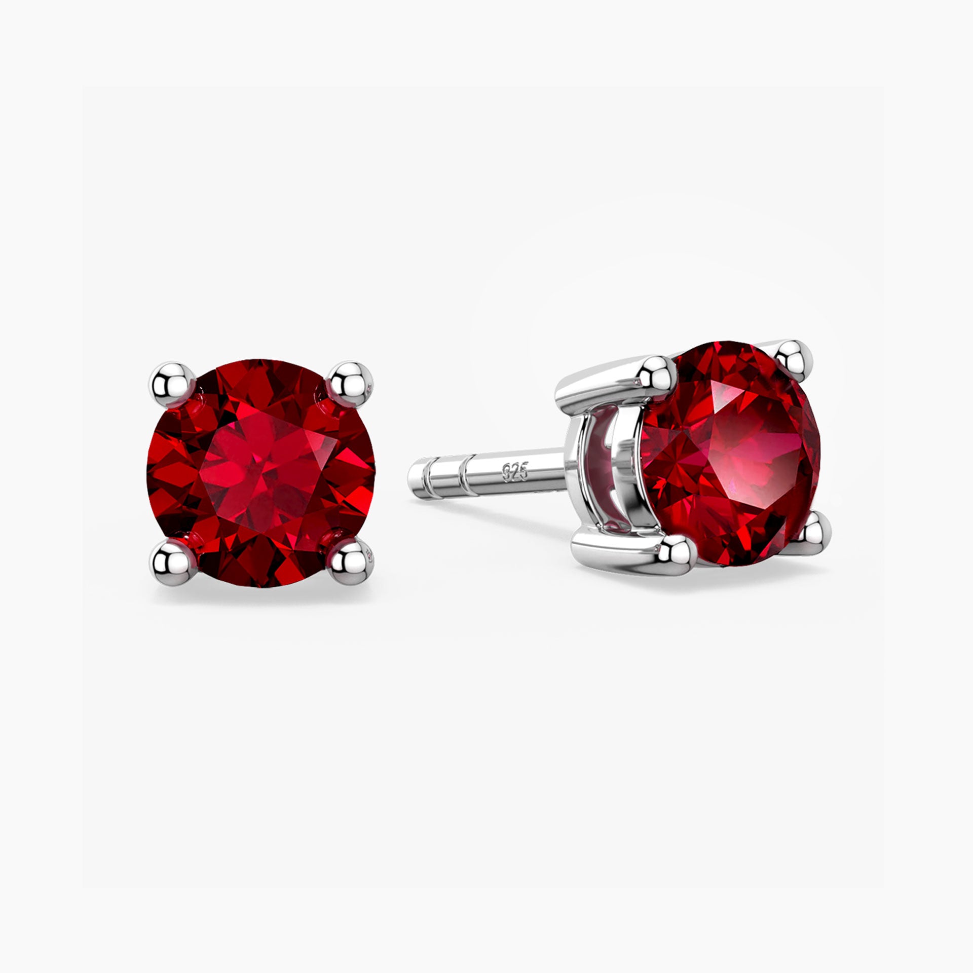 Pair of sterling-silver stud earrings set with round, faceted red rubies—one shown face-on, the other tilted to reveal its four-prong setting and the “S925” hallmark on the post—against a white background.