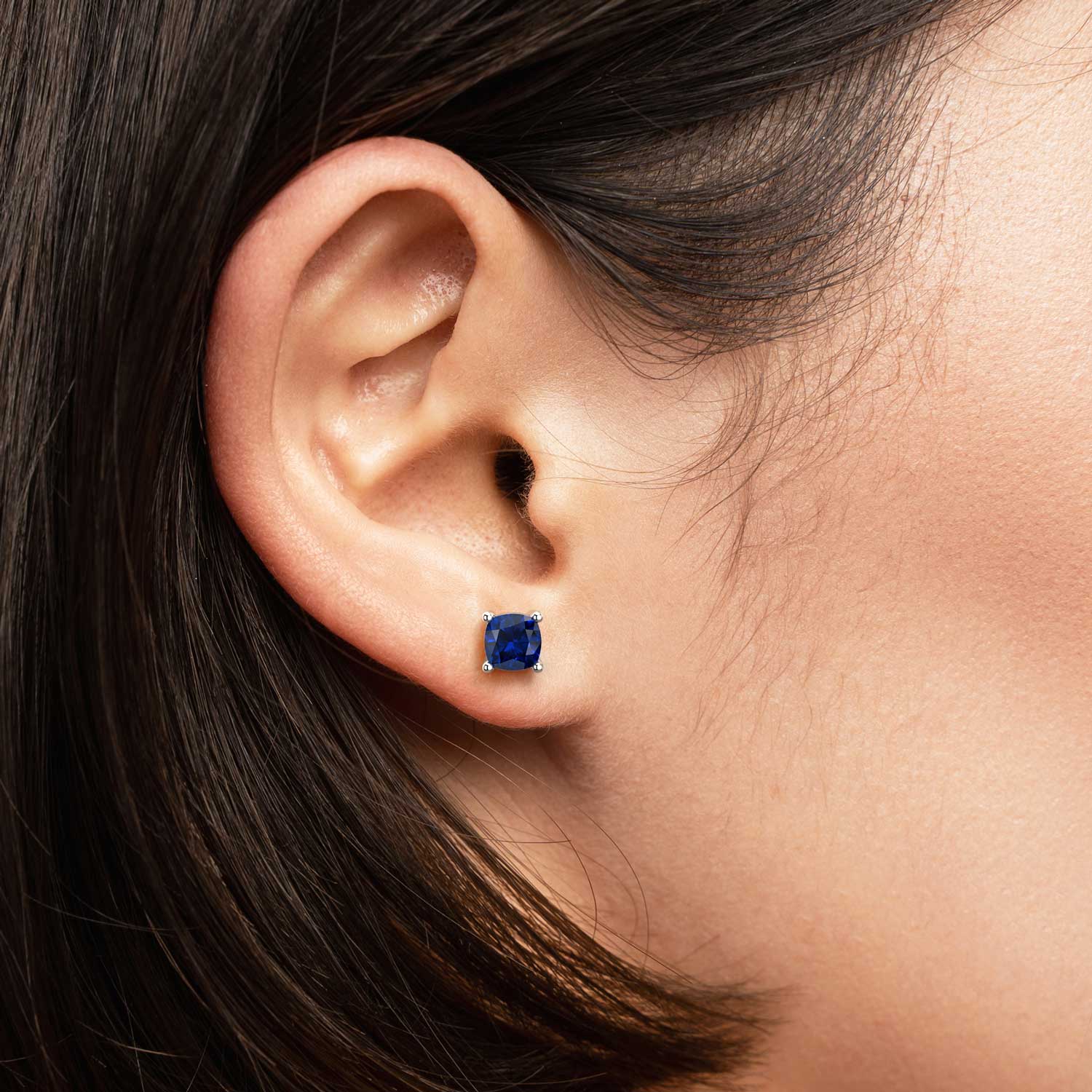 Close-up of a woman’s ear wearing a cushion-cut blue sapphire stud earring in a 925 sterling silver four-prong setting.