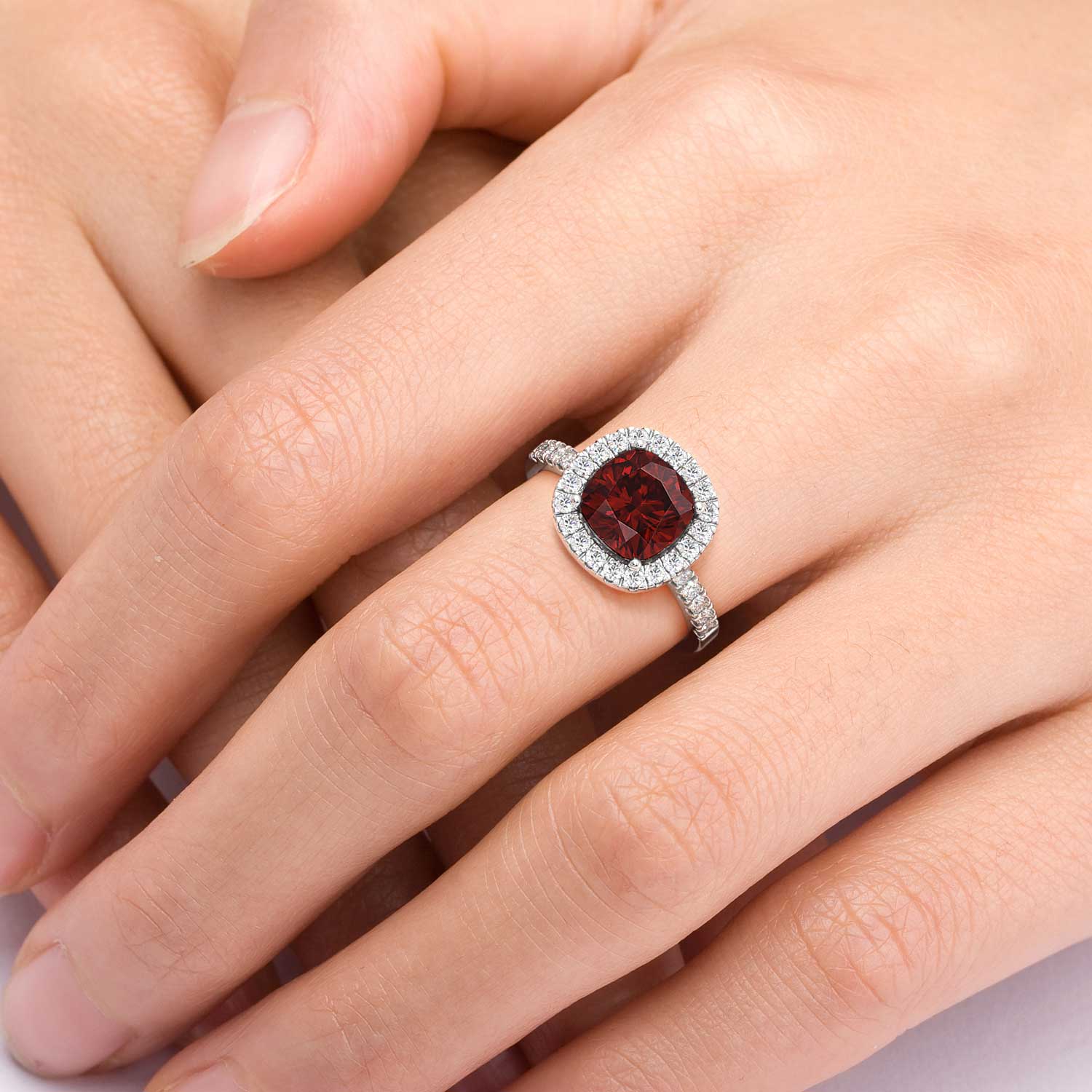 A beautiful cushion-cut garnet halo ring, elegantly displayed on the hand. The deep red hue of the garnet gemstone, surrounded by a halo of shimmering diamonds, adds a touch of sophistication and allure to any ensemble.