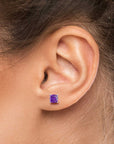 Close-up of a woman’s ear wearing a 6×6 mm cushion-cut amethyst stud earring in a 925 sterling silver four-prong setting.