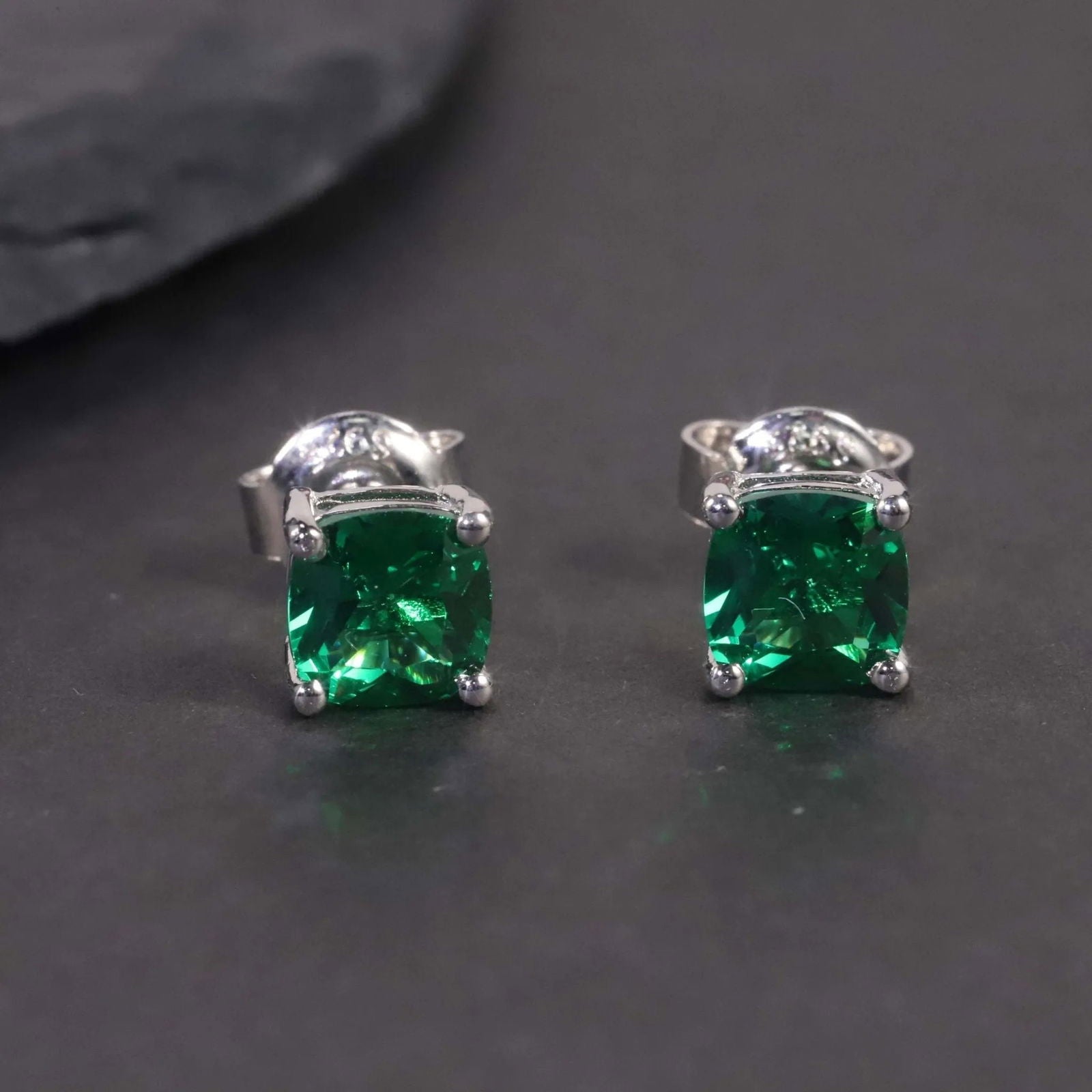 Close-up of the cushion-cut emerald stud earrings displayed on a dark surface.