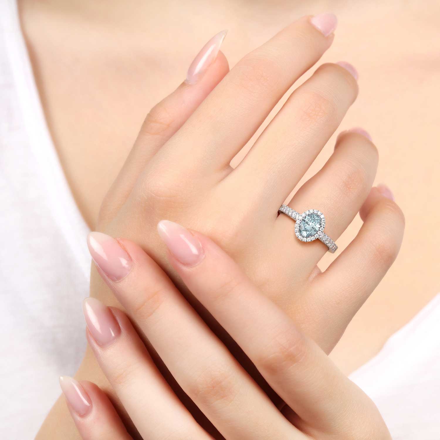 Close-up image of an elegant oval cut aquamarine gemstone ring, set in a halo of sparkling diamonds, worn on a finger.