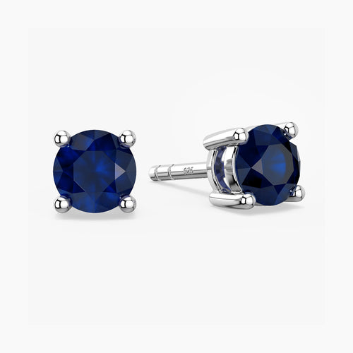 Pair of sterling-silver stud earrings featuring round, faceted deep blue sapphires—one shown front-facing, the other angled to display its four-prong setting and the ‘S925’ hallmark on the post—set against a white background.