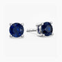 Pair of sterling-silver stud earrings featuring round, faceted deep blue sapphires—one shown front-facing, the other angled to display its four-prong setting and the ‘S925’ hallmark on the post—set against a white background.
