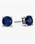 Pair of sterling-silver stud earrings featuring round, faceted deep blue sapphires—one shown front-facing, the other angled to display its four-prong setting and the ‘S925’ hallmark on the post—set against a white background.