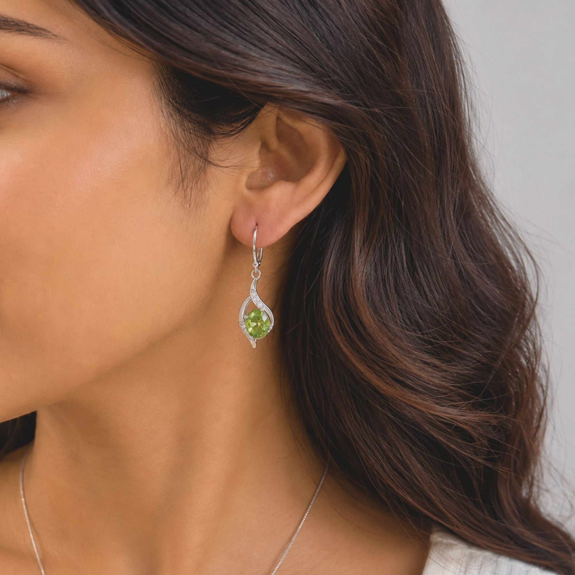 A close-up of a woman wearing a silver peridot dangle earring with a green peridot gemstone and clear accent stones, shown against her ear and dark brown hair.