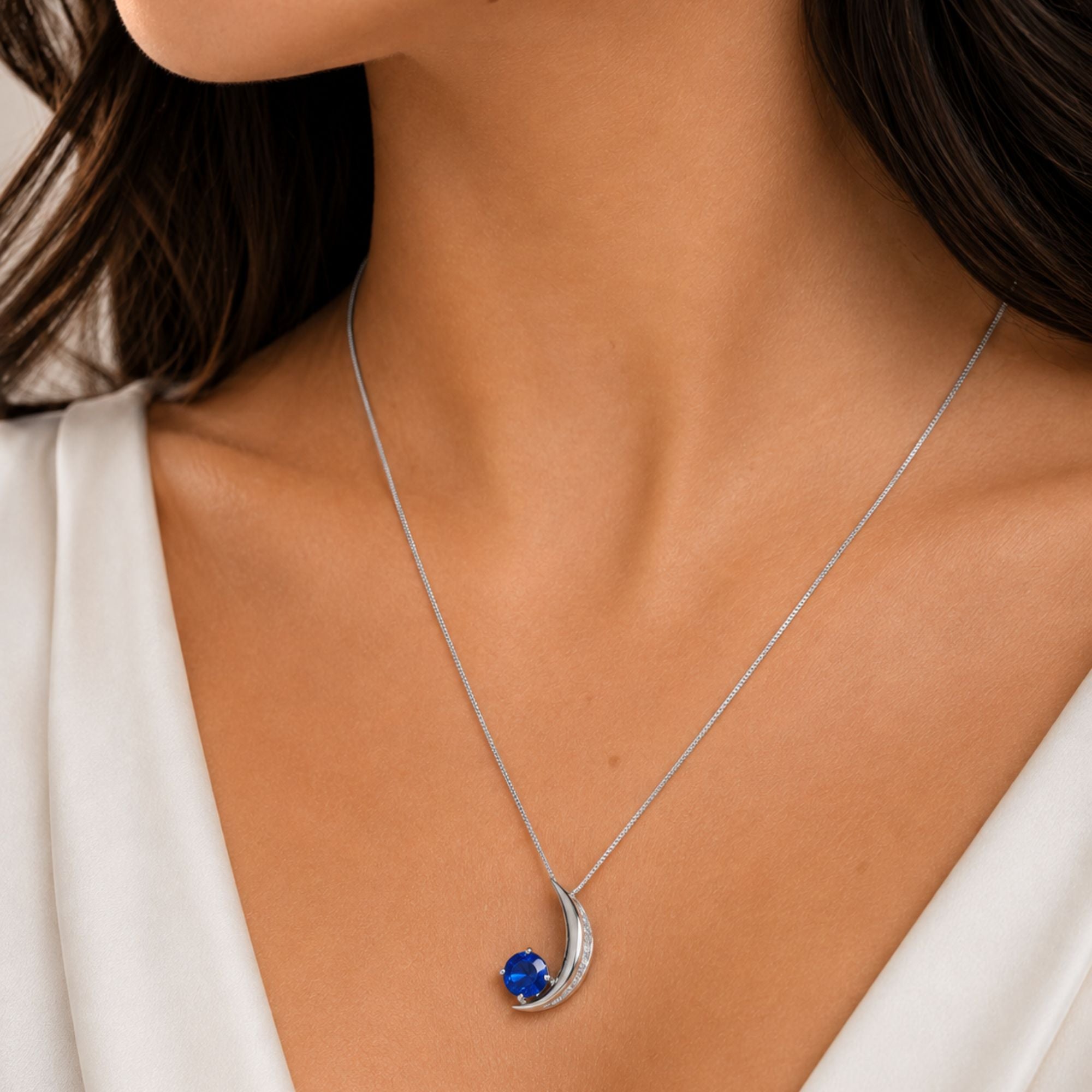 A close-up of a model wearing a silver crescent sapphire moon pendant necklace with a vibrant blue gemstone, styled with a white top.