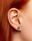 Close-up of a red-haired woman’s ear wearing a cushion-cut London blue topaz stud earring set in 925 sterling silver.