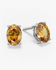 Pair of oval-cut golden yellow citrine stud earrings in a four-prong silver setting with push-back closures, displayed on a white background