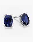 Pair of oval-cut deep blue sapphire stud earrings in a four-prong silver setting with push-back closures, displayed on a white background