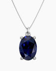 Oval-cut deep blue sapphire pendant in a four-prong silver setting, suspended from a fine silver chain, displayed on a white background.