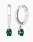 Close-up of silver hoop earrings with oval emerald drops, showcasing their polished finish and vibrant green gemstones.
