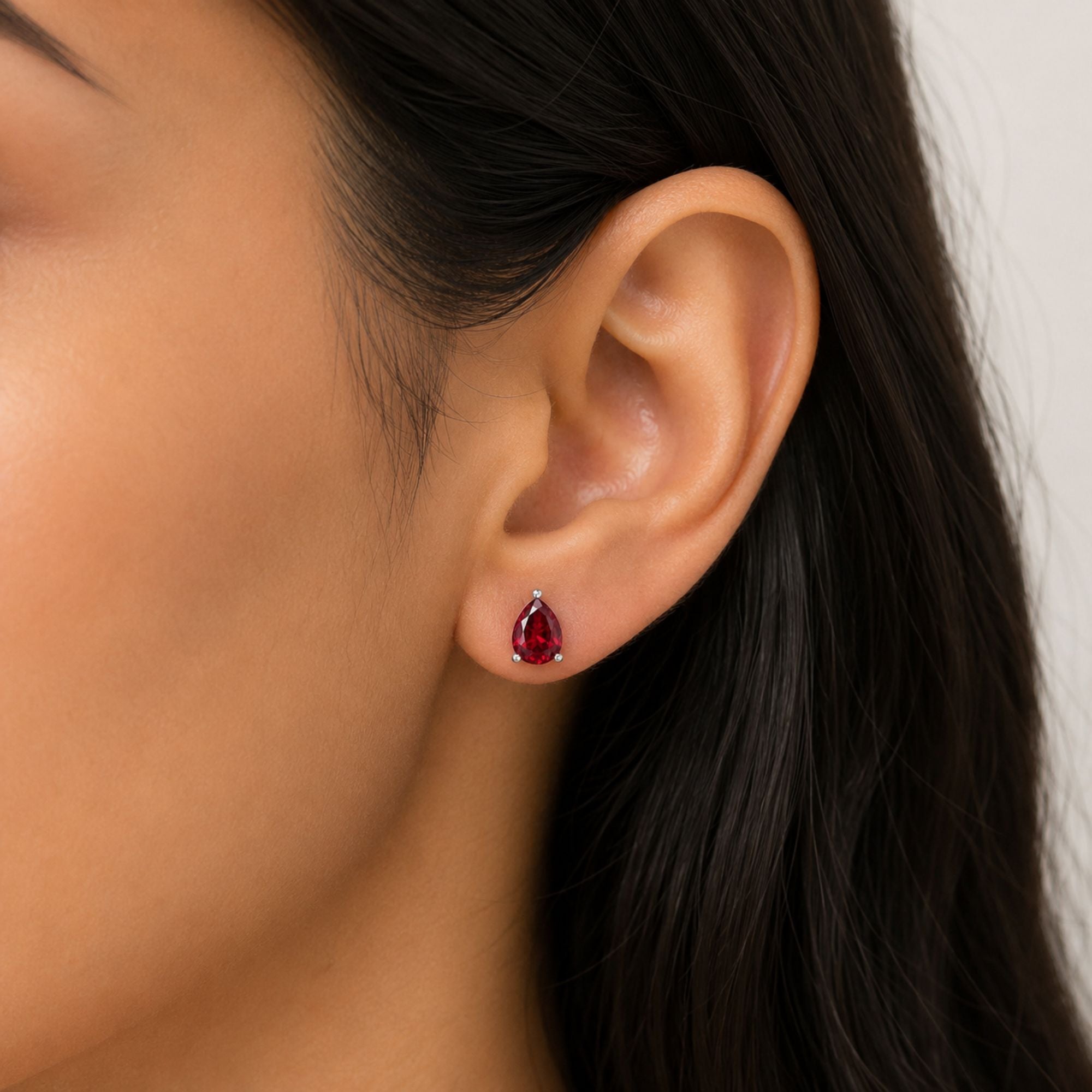A close-up of a model's ear wearing a small teardrop-shaped red gemstone stud earring with a silver-toned setting.