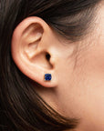 Close-up of a woman’s ear wearing a cushion-cut blue sapphire stud earring in a 925 sterling silver four-prong setting.