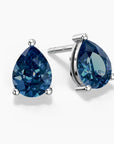 Two pear-shaped London blue topaz stud earrings in sterling silver three-prong settings—one shown from the front and the other at an angle—featuring rich deep-blue gemstones with brilliant faceting, displayed on a white background.
