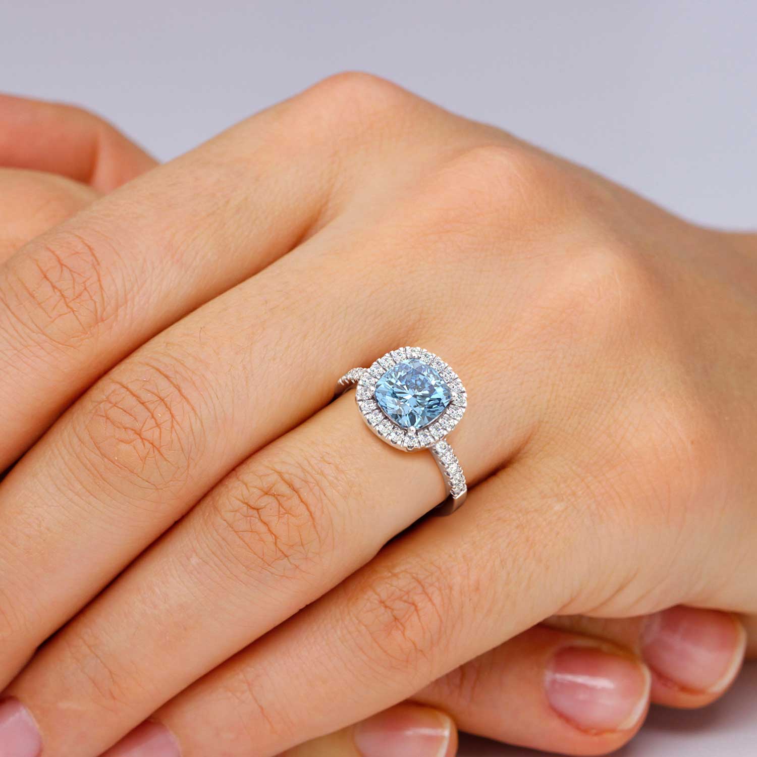 A breathtaking cushion-cut topaz halo ring, elegantly displayed on the hand. The mesmerizing blue hue of the topaz gemstone, accentuated by a shimmering halo of diamonds, exudes sophistication and charm, creating a striking piece of jewelry.